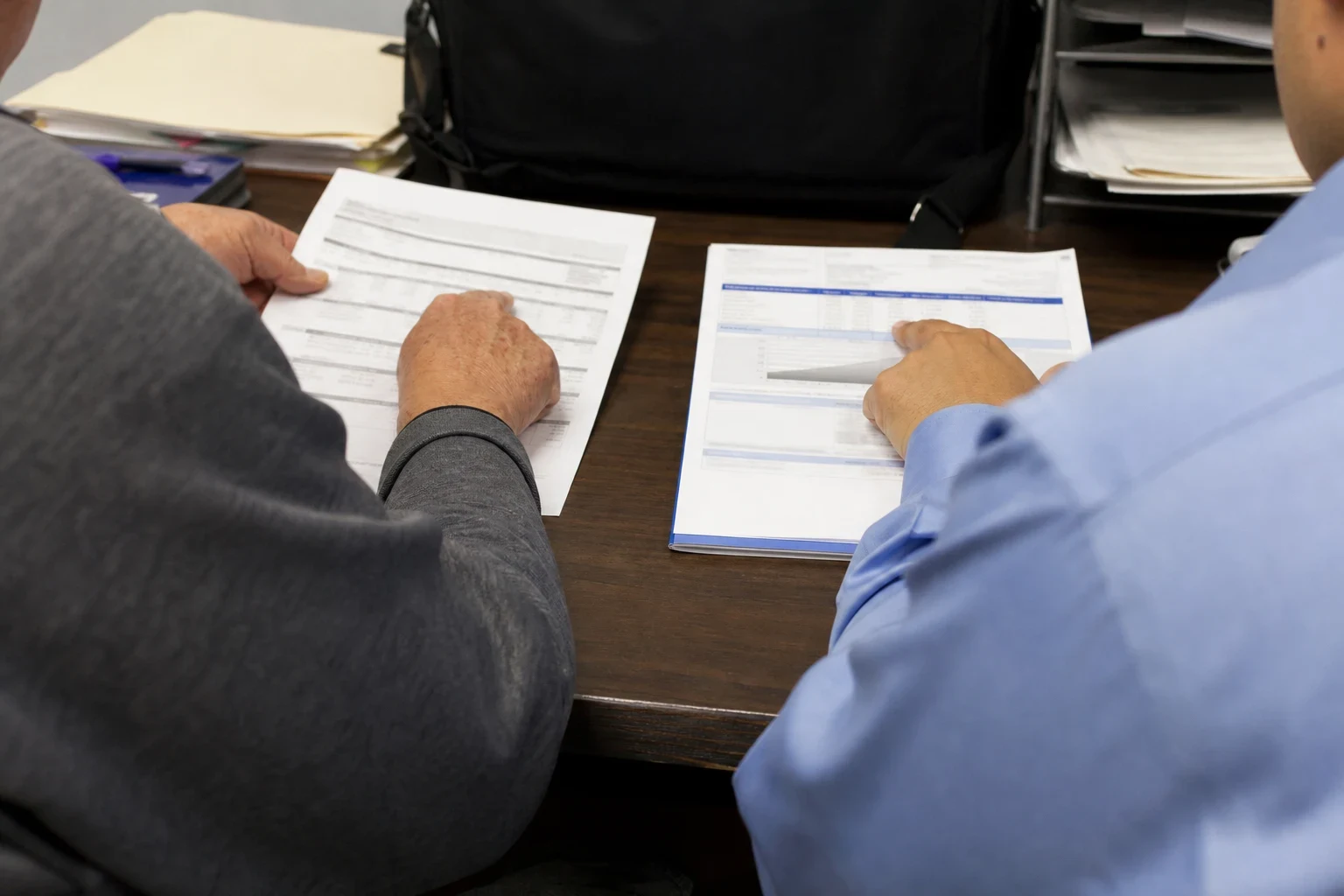 Over-the-shoulder view of two people at a wooden office desk comparing a printed statement and a coverage document laid side by side, with folders and a paper tray behind.