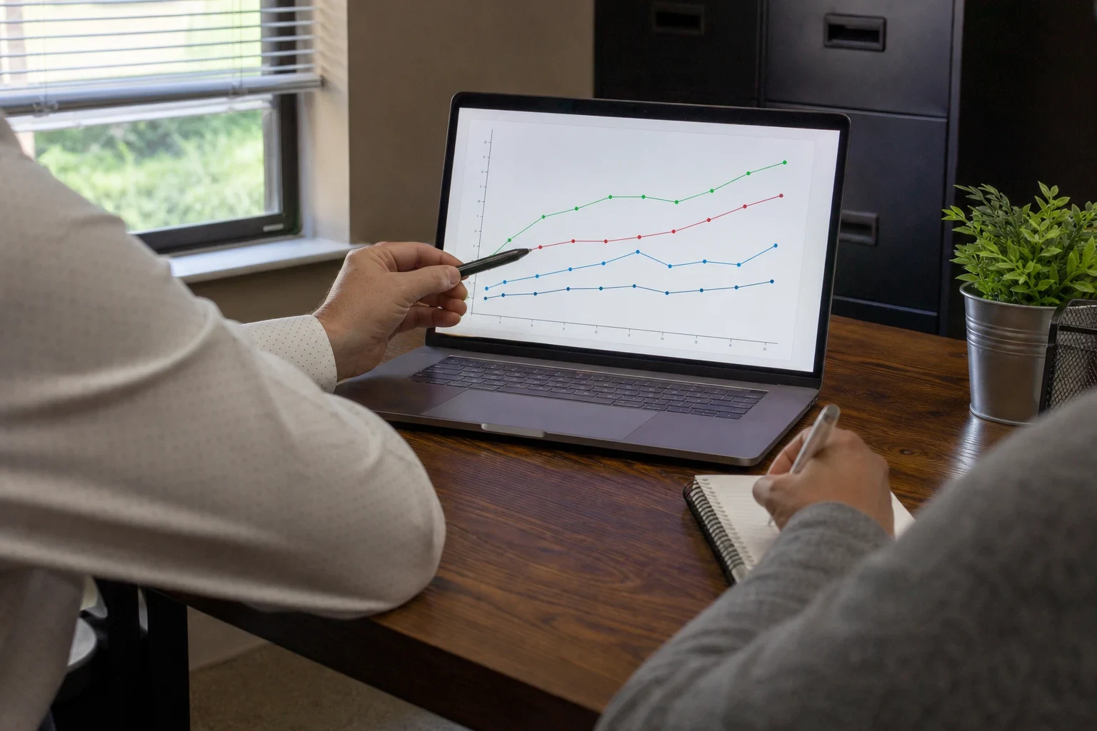 Agent pointing a pen at a line chart on a laptop at a wooden desk while a client takes notes in a spiral notebook, filing cabinet and window behind